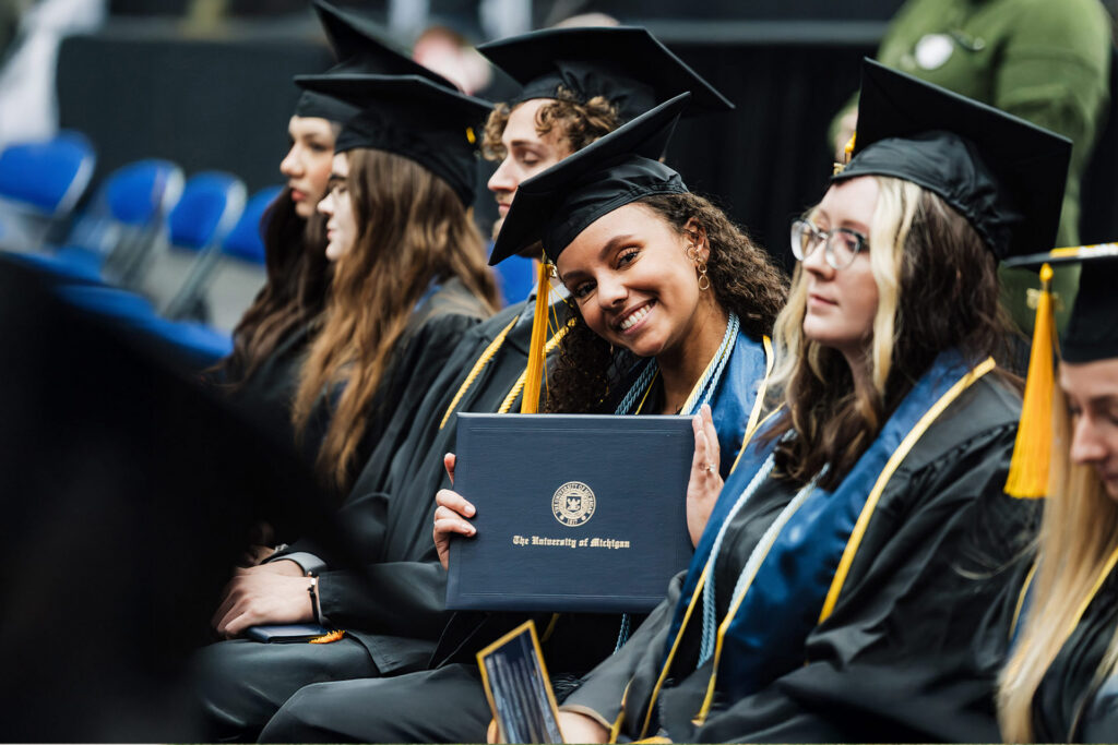 A group of graduates in caps and gowns sitting together, with one holding a diploma from The University of Michigan.