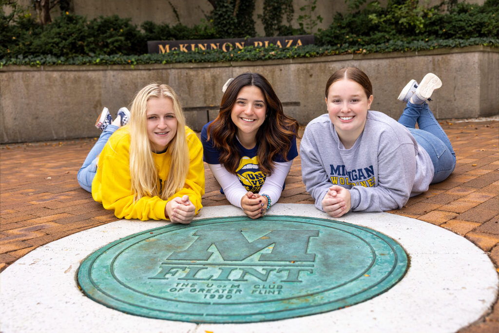 Three individuals are lying on the ground, posing with their hands clasped in front of a circular emblem that reads "M FLINT" in the center. The background features greenery and a sign that says "McKinnon Plaza."