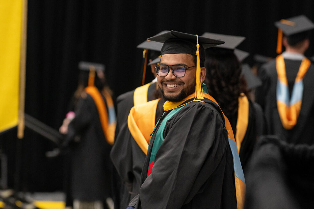 A graduate in a black cap and gown stands among fellow graduates, wearing a colorful stole and tassel, in a graduation ceremony setting.