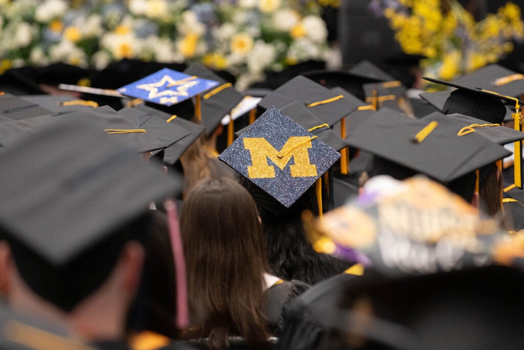 A crowd of graduates wearing black caps and gowns, with one cap prominently displaying a glittery gold "M" on a blue background.