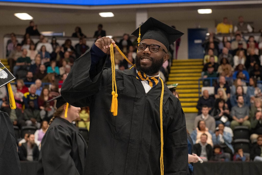 A graduate in a black cap and gown holds up a yellow tassel, celebrating at a graduation ceremony with an audience in the background.