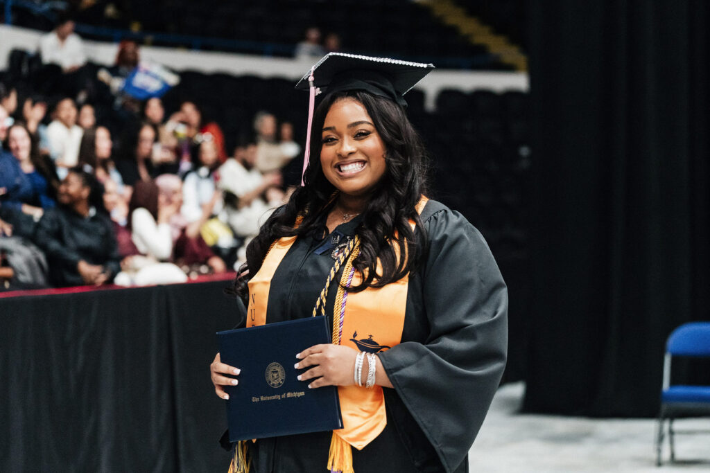 A graduate wearing a black cap and gown, holding a diploma from the University of Michigan, stands in front of a crowd at a graduation ceremony.