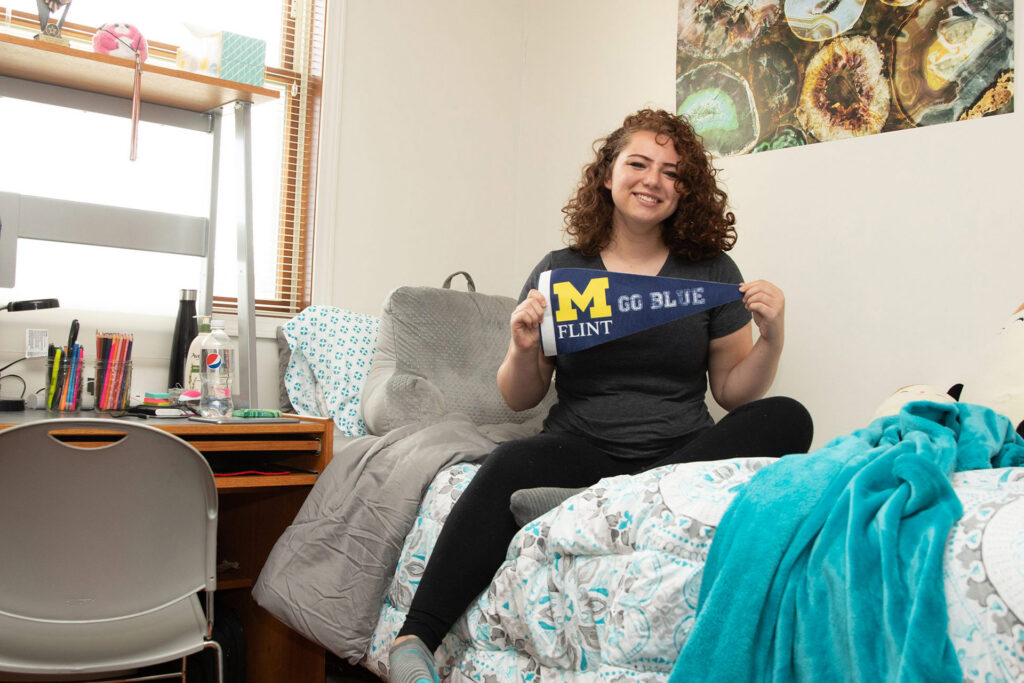 A person sits on a bed in a dorm room, holding a pennant that says "M FLINT GO BLUE." A desk with school supplies is visible.