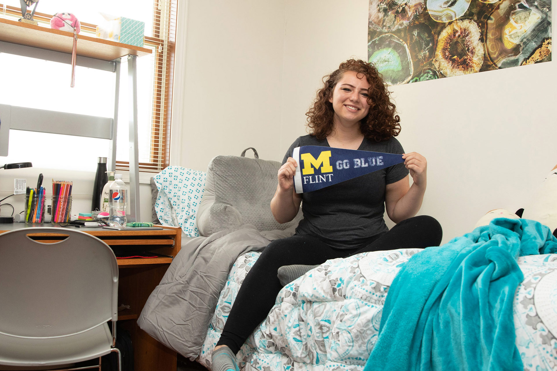 A person sits on a bed in a dorm room, holding a pennant that says "M FLINT GO BLUE." A desk with school supplies is visible.