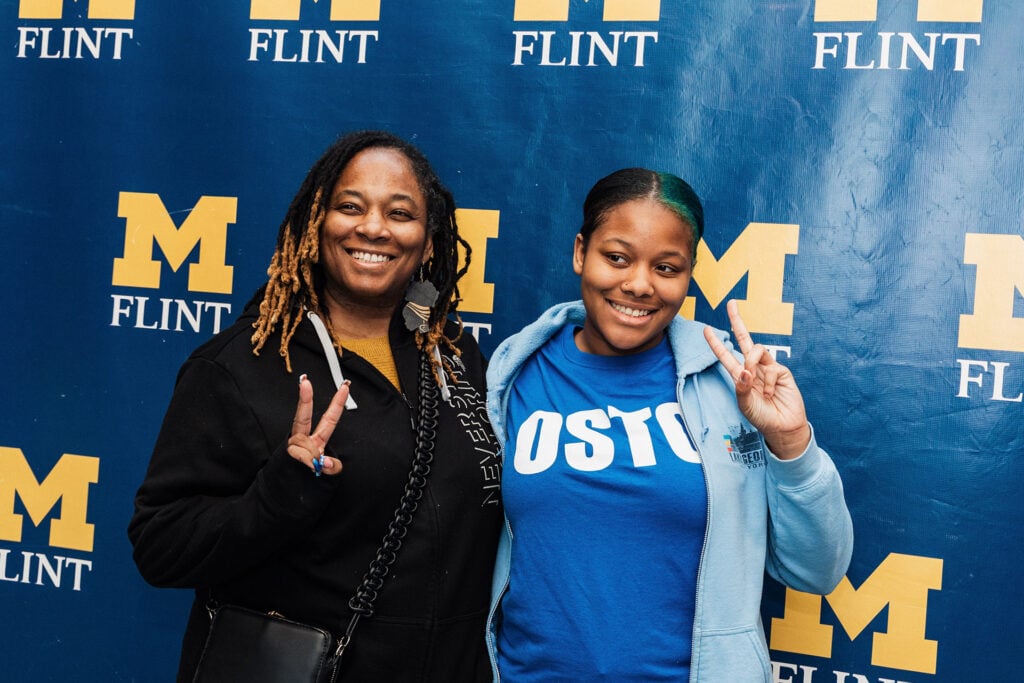 Two people pose together, each making a peace sign, in front of a blue backdrop with "M FLINT" displayed prominently.