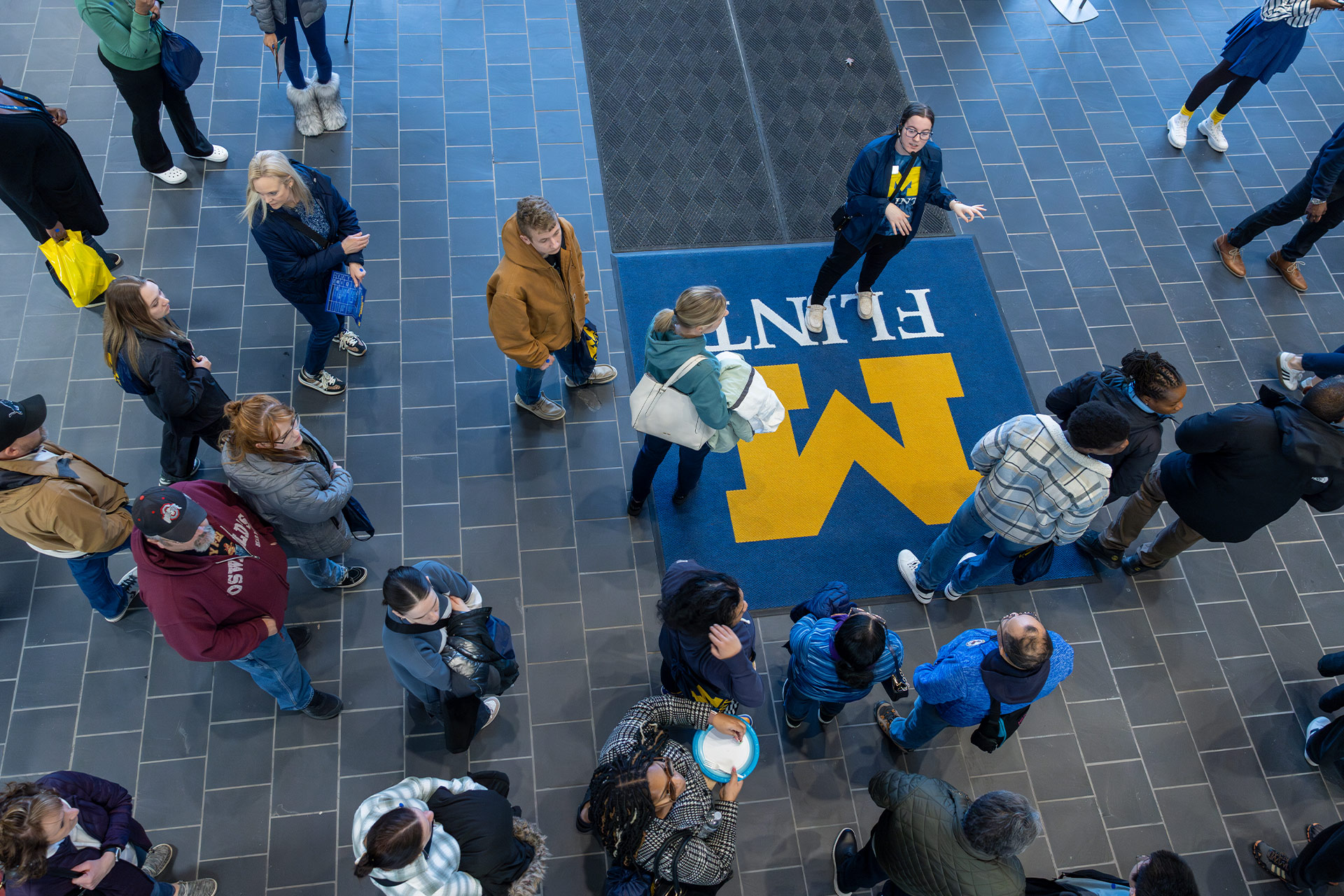 Aerial view of a crowd of people gathered in a lobby area with a large blue and yellow logo on the floor.
