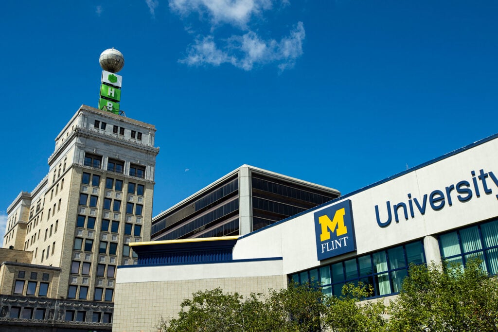 A historic building with a green sign and a globe on top, next to a modern building labeled "University of Michigan Flint."