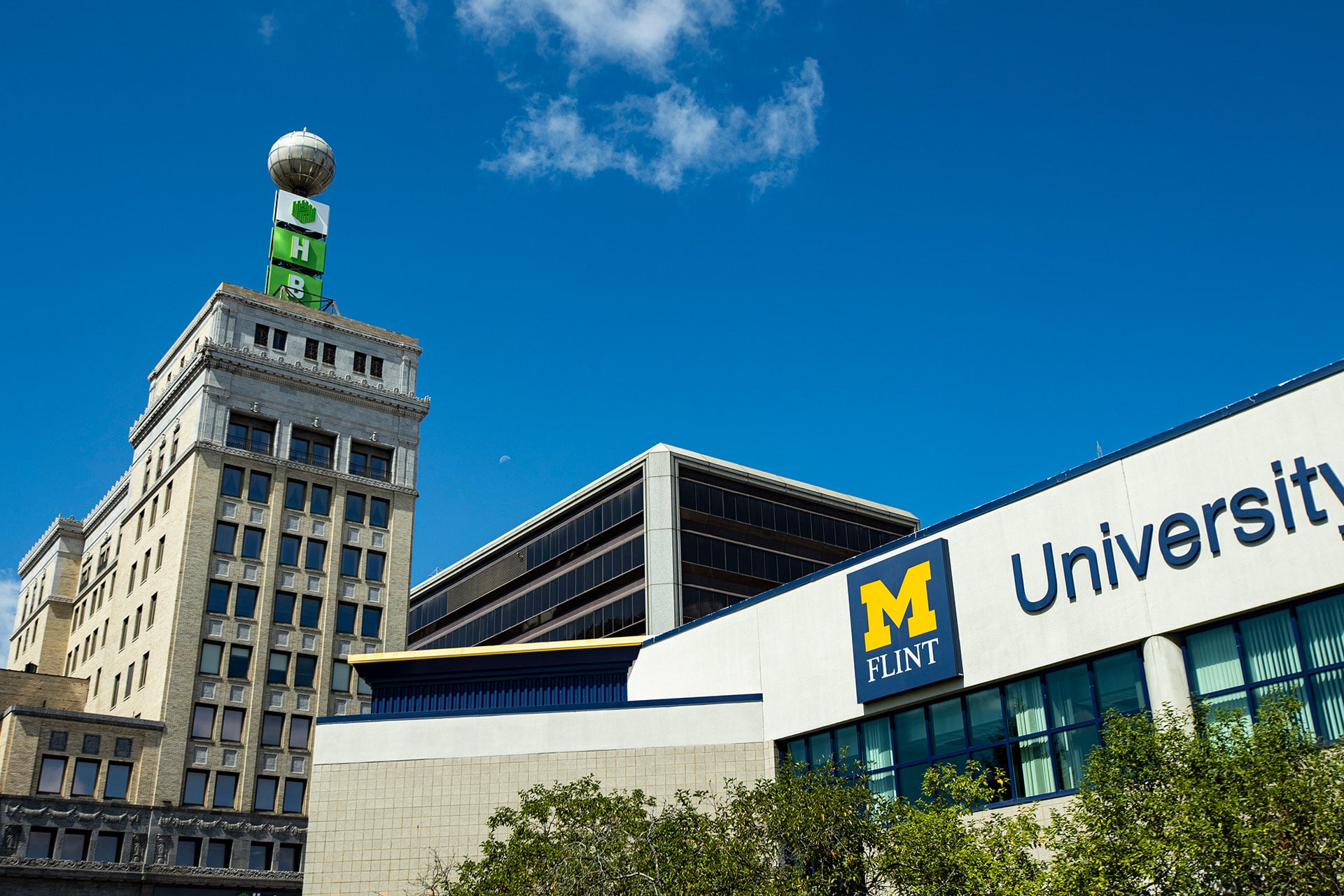 A historic building with a green sign and a globe on top, next to a modern building labeled "University of Michigan Flint."