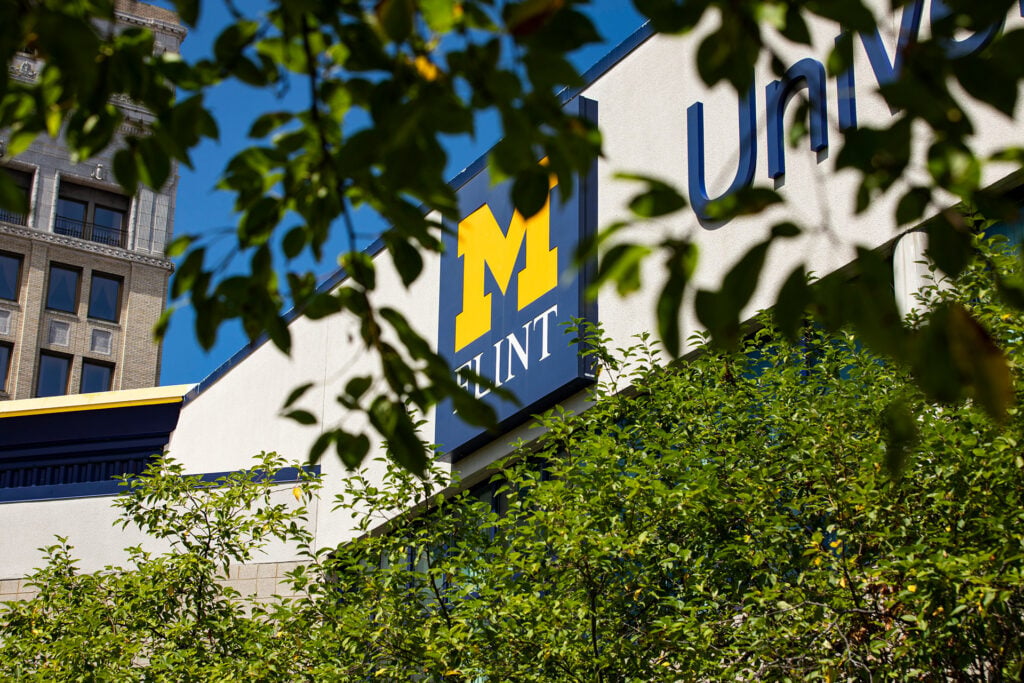Sign for the University of Michigan-Flint partially obscured by green leaves, with a building in the background.