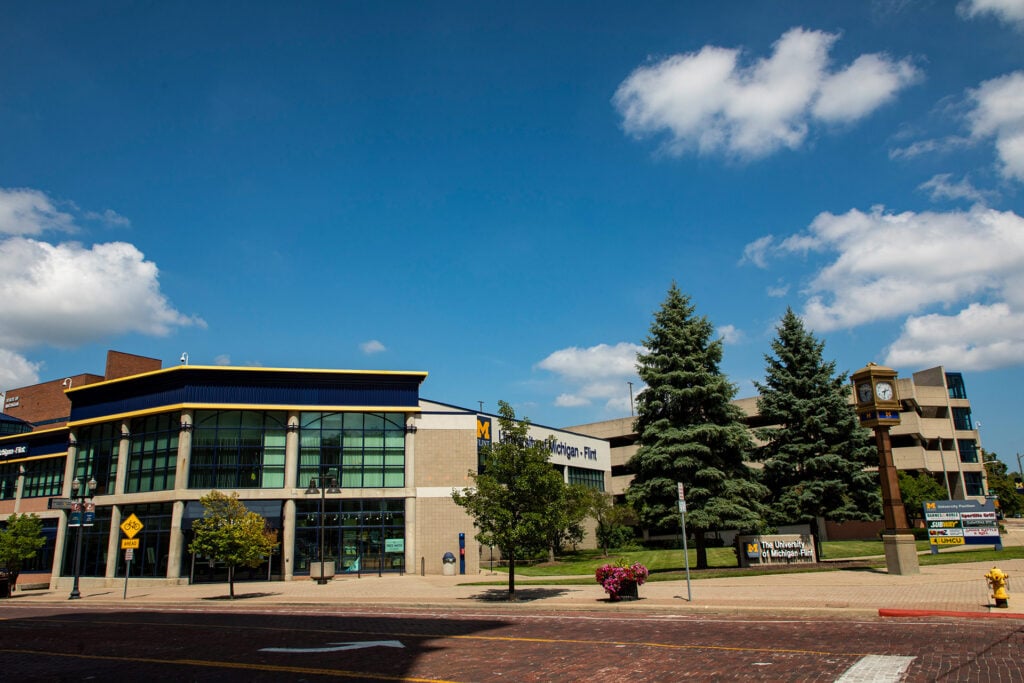 A view of a university building with large glass windows, flanked by trees and a clock tower, under a blue sky with clouds.