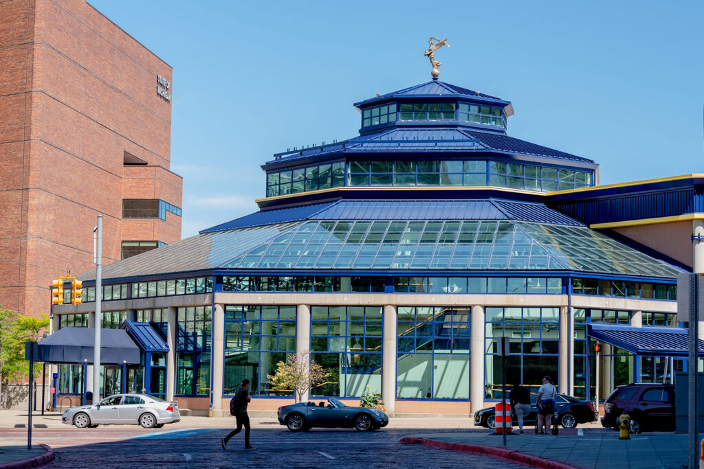 A modern glass building with a blue roof and a statue on top, surrounded by cars and pedestrians on a sunny day.