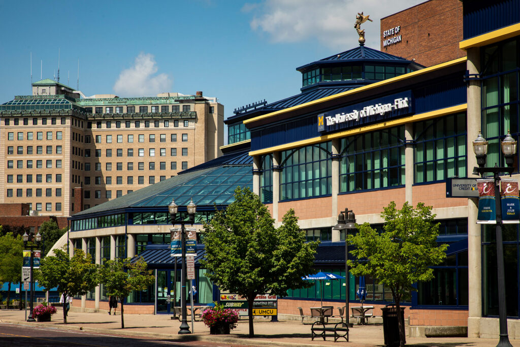 The image shows the University of Michigan-Flint building with a glass roof and surrounding trees, set against a blue sky.