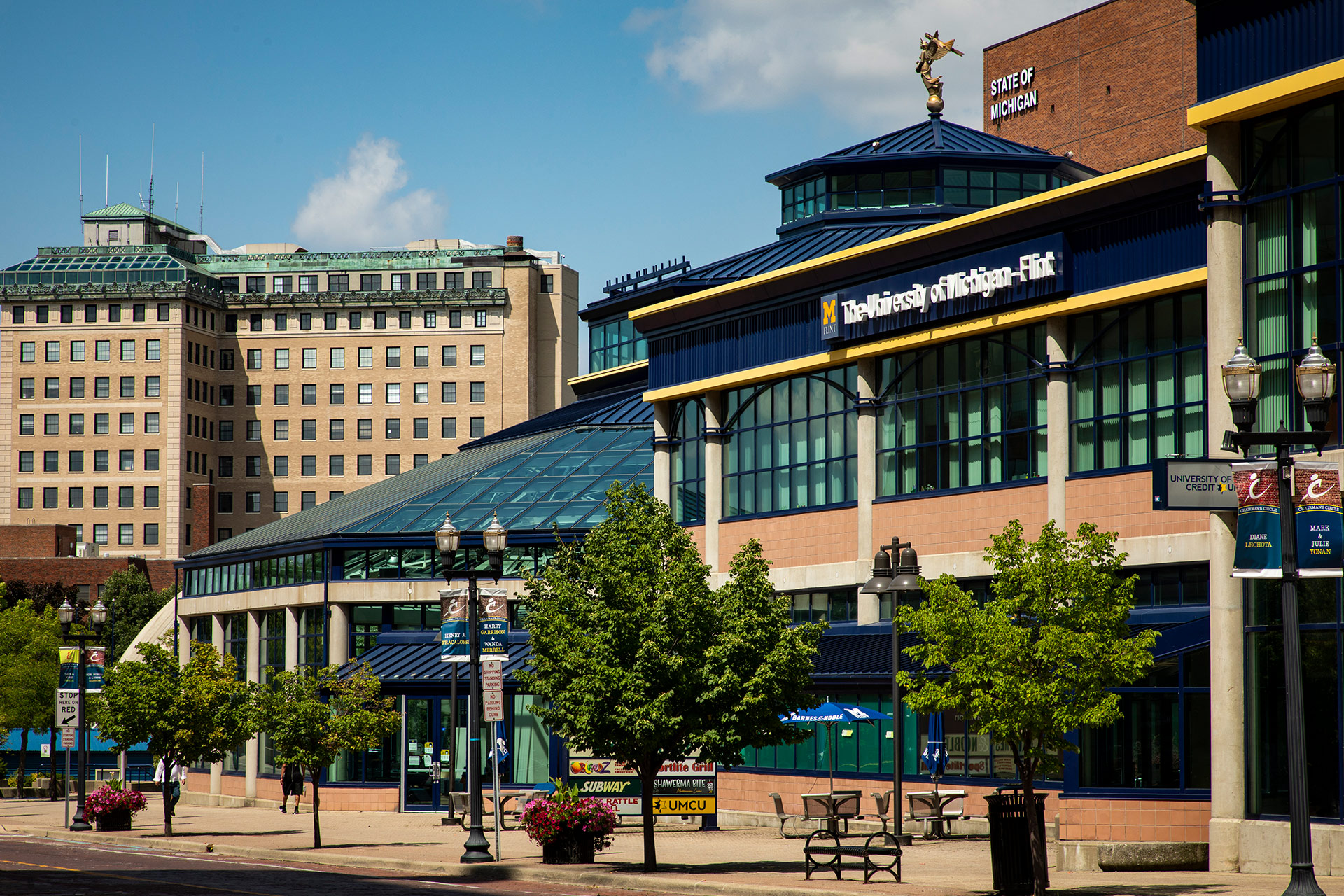The image shows the University of Michigan-Flint building with a glass roof and surrounding trees, set against a blue sky.
