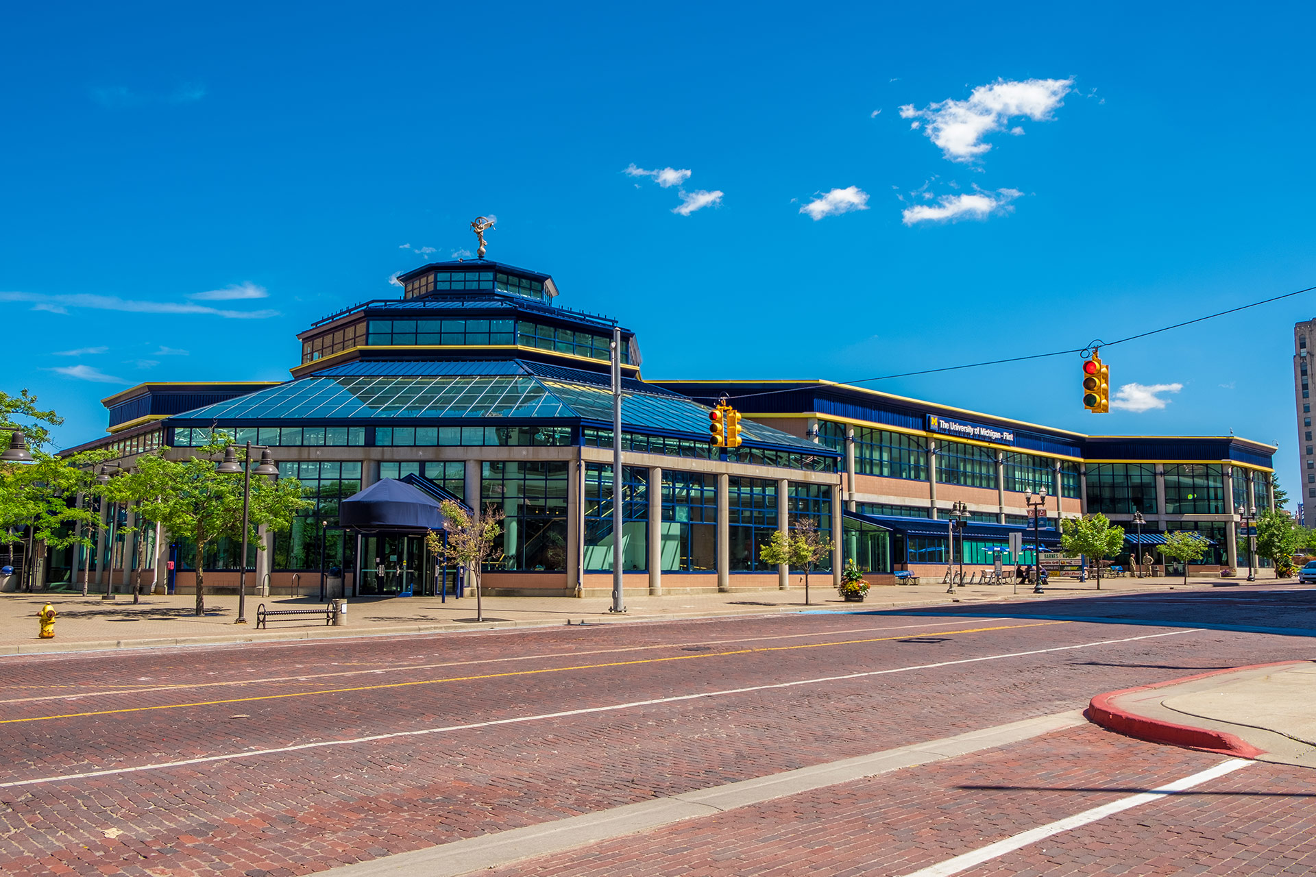 A large, modern building with a glass roof and a statue on top, surrounded by trees and a brick-paved street under a blue sky.