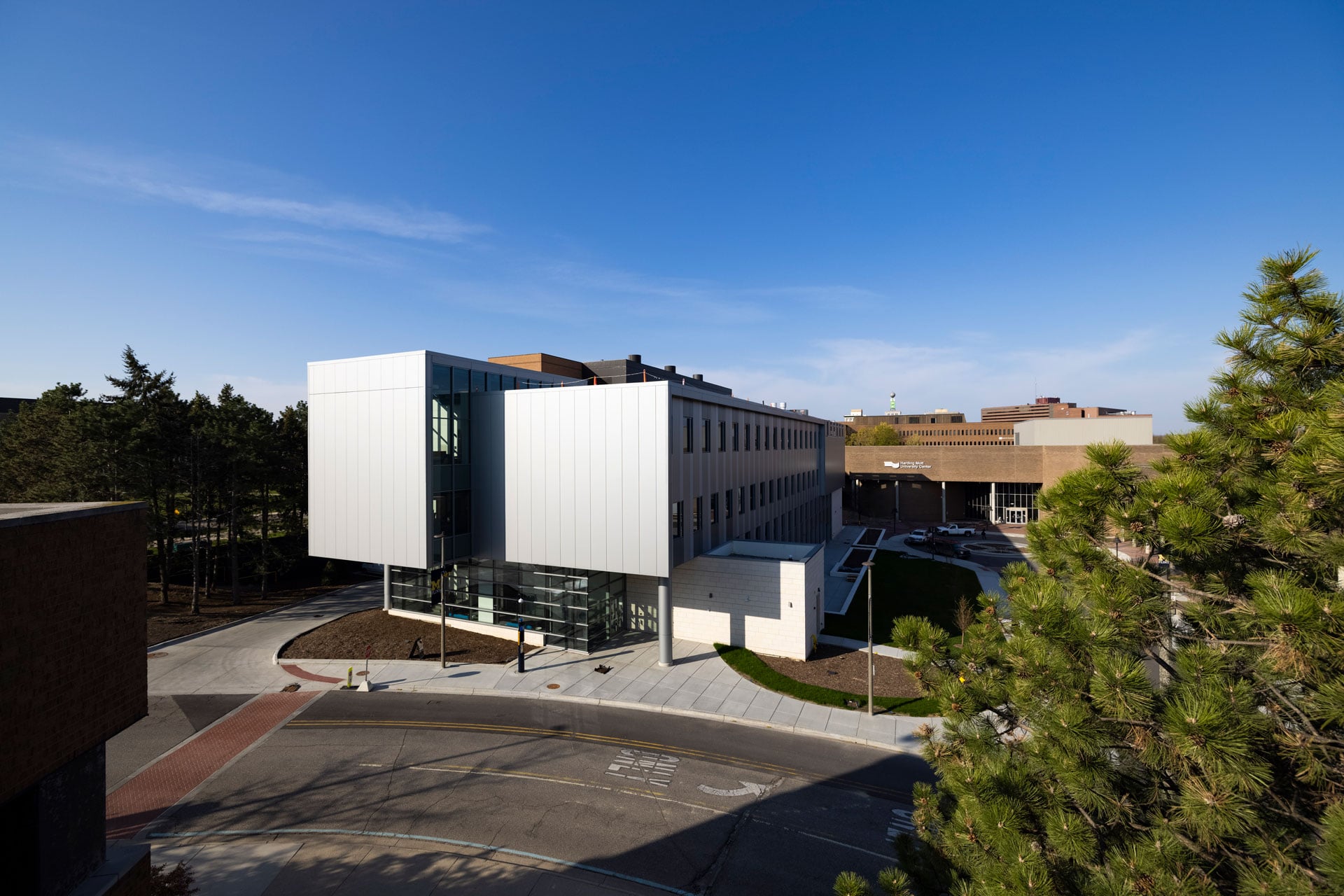 A modern building with a sleek, silver facade is shown, surrounded by trees and a clear blue sky.
