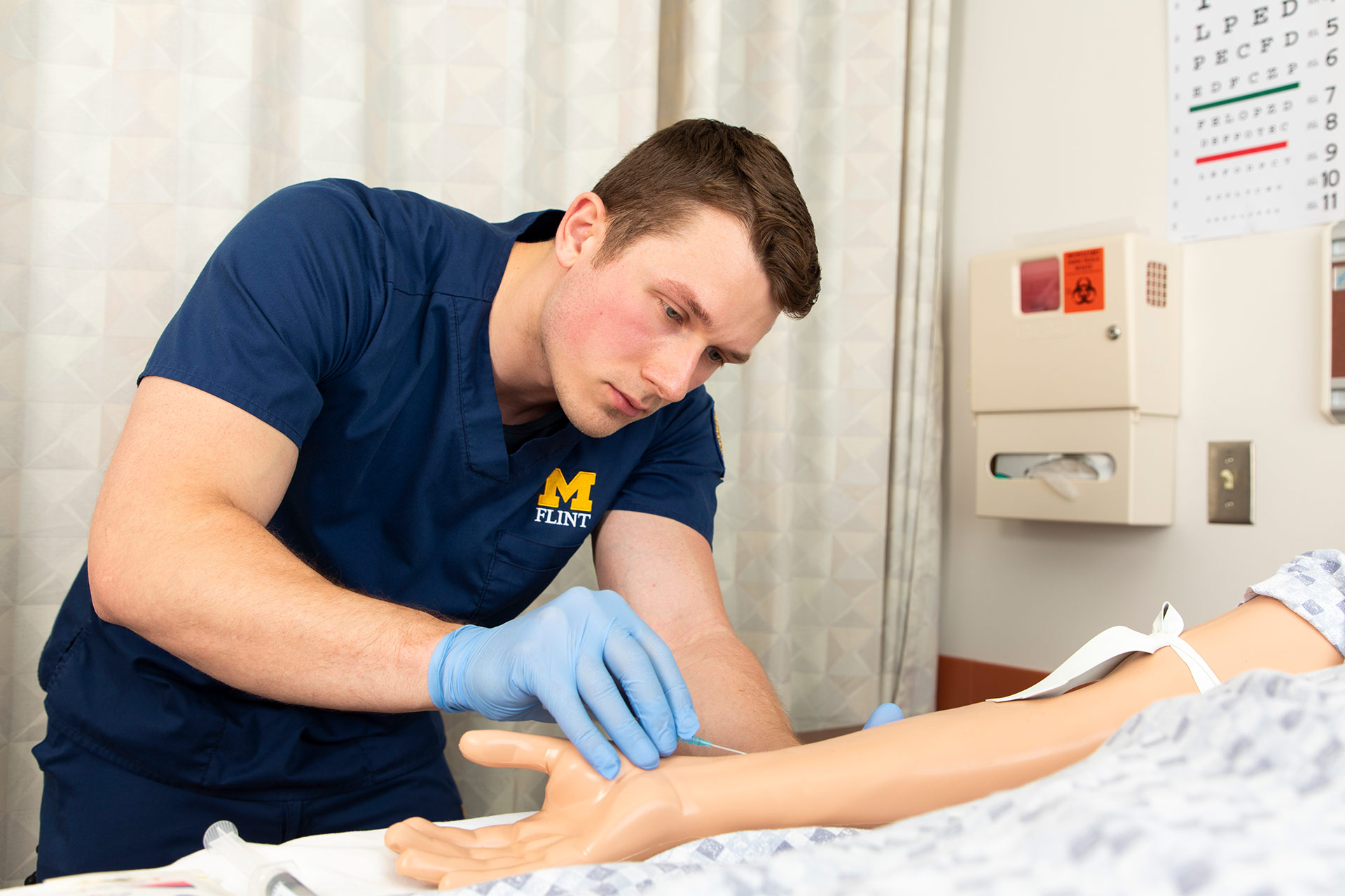 A person in scrubs is practicing a medical procedure on a training mannequin's arm in a clinical setting.