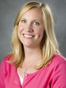 Rachel Gutierrez smiles while wearing a pink, buttoned sweater and a silver necklace. Her medium-length, blonde hair is hanging down onto her shoulders.