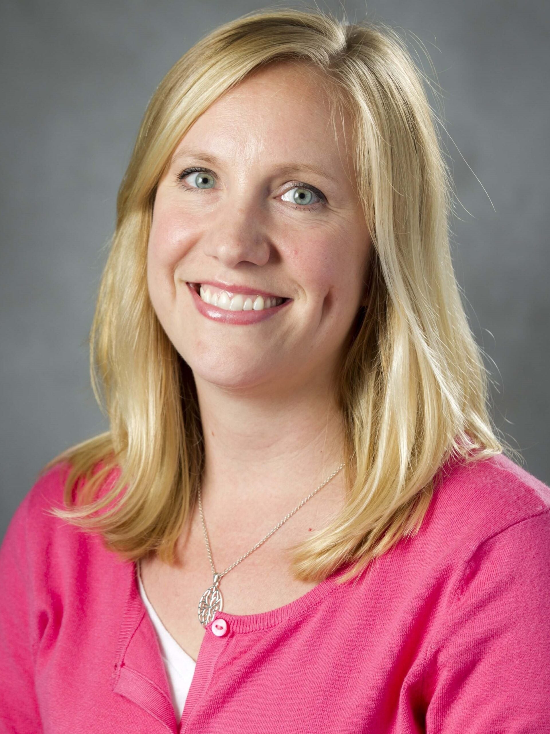Rachel Gutierrez smiles while wearing a pink, buttoned sweater and a silver necklace. Her medium-length, blonde hair is hanging down onto her shoulders.