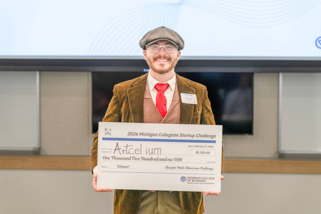 A young man wearing a brown corduroy jacket, red tie and flat cap holding a large check for $1,500 made out to "Artcelium," winner of the 2026 Michigan Collegiate Startup Challenge.