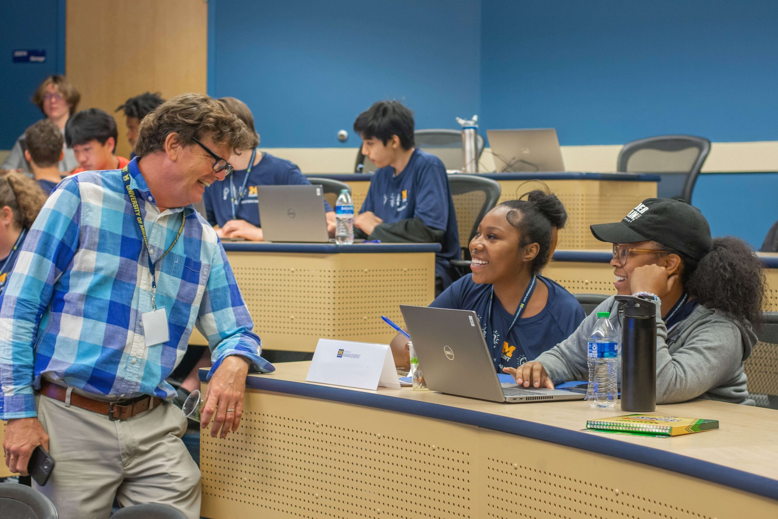 A professor in a checkered shirt interacts with two students. Both students are seated at a classroom desk with a laptop during the Summer Entrepreneurship Institute.
