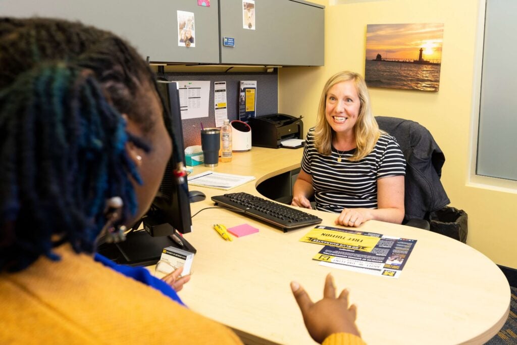 A woman sits at a desk smiling while speaking to a student. University of Michigan-Flint literature sits on the desk between them.