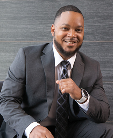 A professional portrait of a smiling man with a black beard and hair. He is seated and wearing a charcoal-gray suit, black tie with a diamond pattern and a smart watch. Behind him is a wood-grained, paneled wall that is grayish brown.