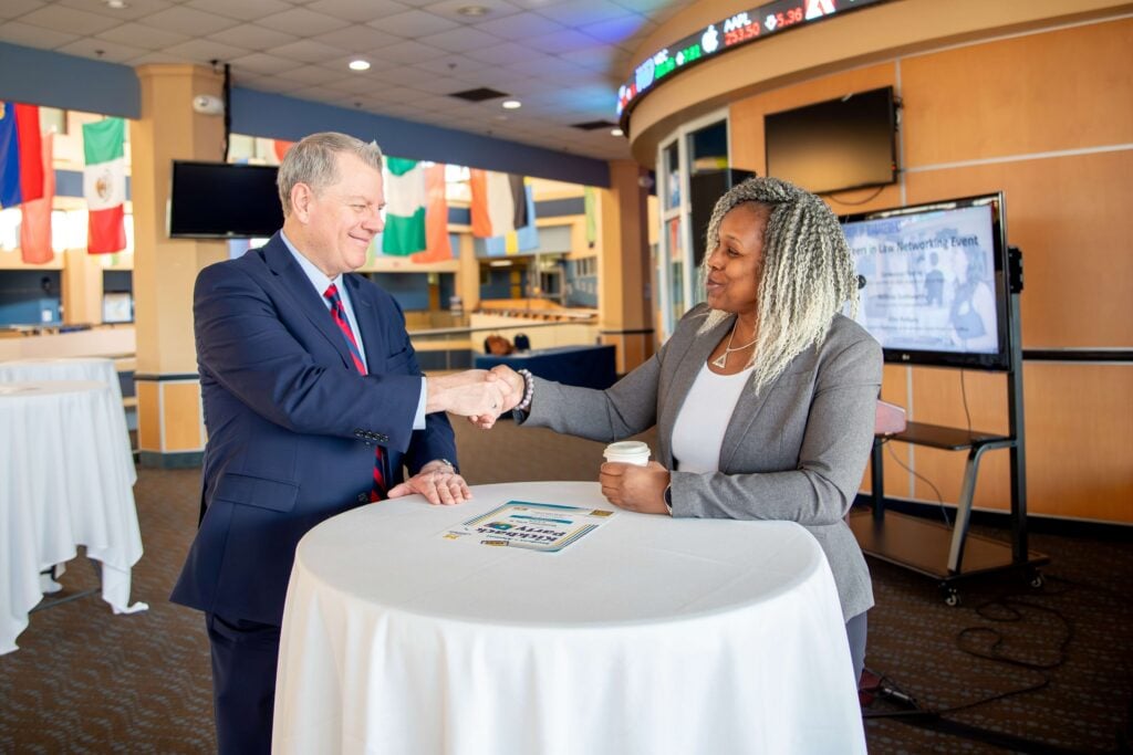 A man and woman shake hands at a business networking event. Flags and a presentation screen appear in the background.