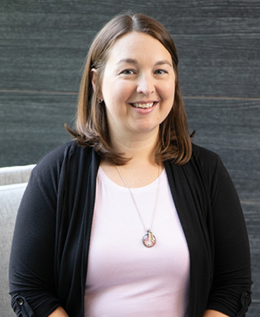 A portrait of a smiling woman with shoulder-length brown hair. She is wearing a light-pink shirt and a black cardigan with a decorative pendant necklace. Behind her is a wood-grained, paneled wall that is grayish brown.
