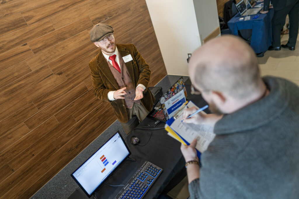 A young, bearded man gestures while speaking to another person who is taking notes at a table with a computer. The young man is wearing a brown corduroy jacket, brown flat cap and red tie.