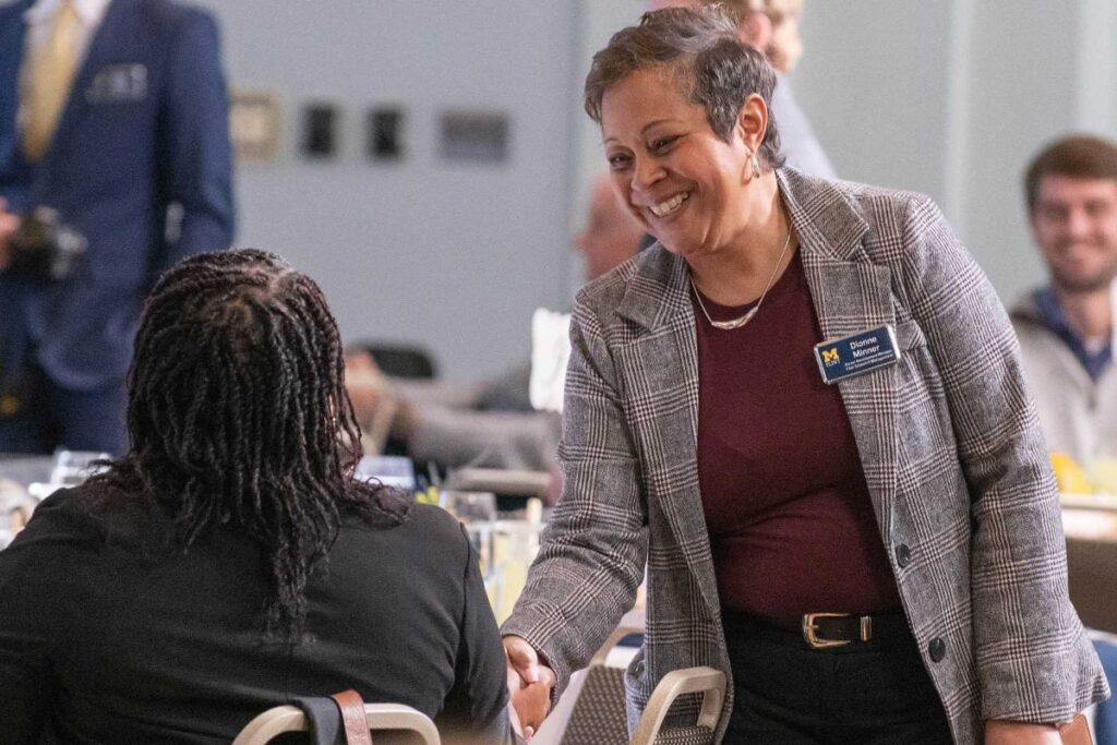 A woman smiles and shakes hands with a student who is seated at a business event. Tables are set for lunch in the background.