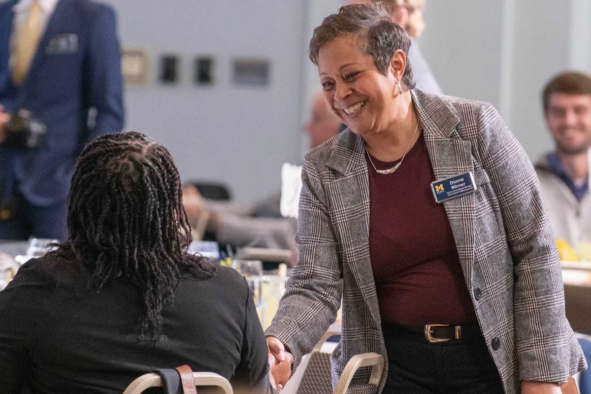 A woman smiles and shakes hands with a student seated at a business event. Tables are set for lunch in the background.