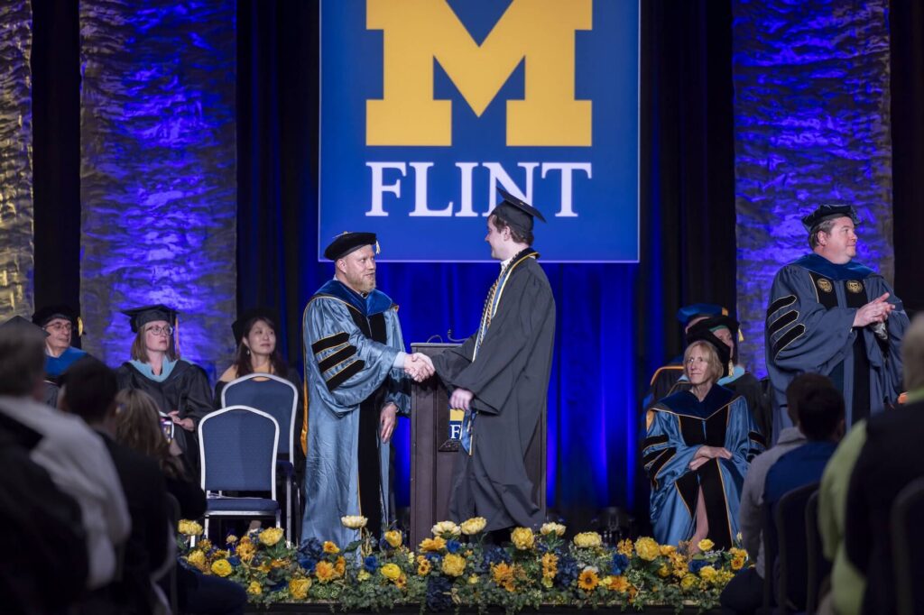 A graduate shakes hands with a faculty member on stage during a graduation ceremony. A large University of Michigan-Flint banner is hanging in the background.