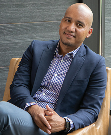 A bald man seated in a light-brown, leather chair, looking relaxed. He is wearing a navy-blue blazer, a blue-and-white, plaid shirt, and light-blue pants. Behind him is a wood-grained, paneled wall that is grayish brown.