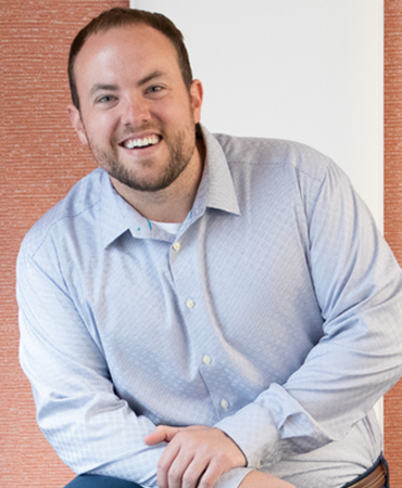 A professional portrait of a man with receding brown hair and a short beard who is looking directly at the camera. He is wearing a light-blue, button-down shirt and navy-blue pants. The background is a vertical, woven tapestry that is light red on an off-white wall.