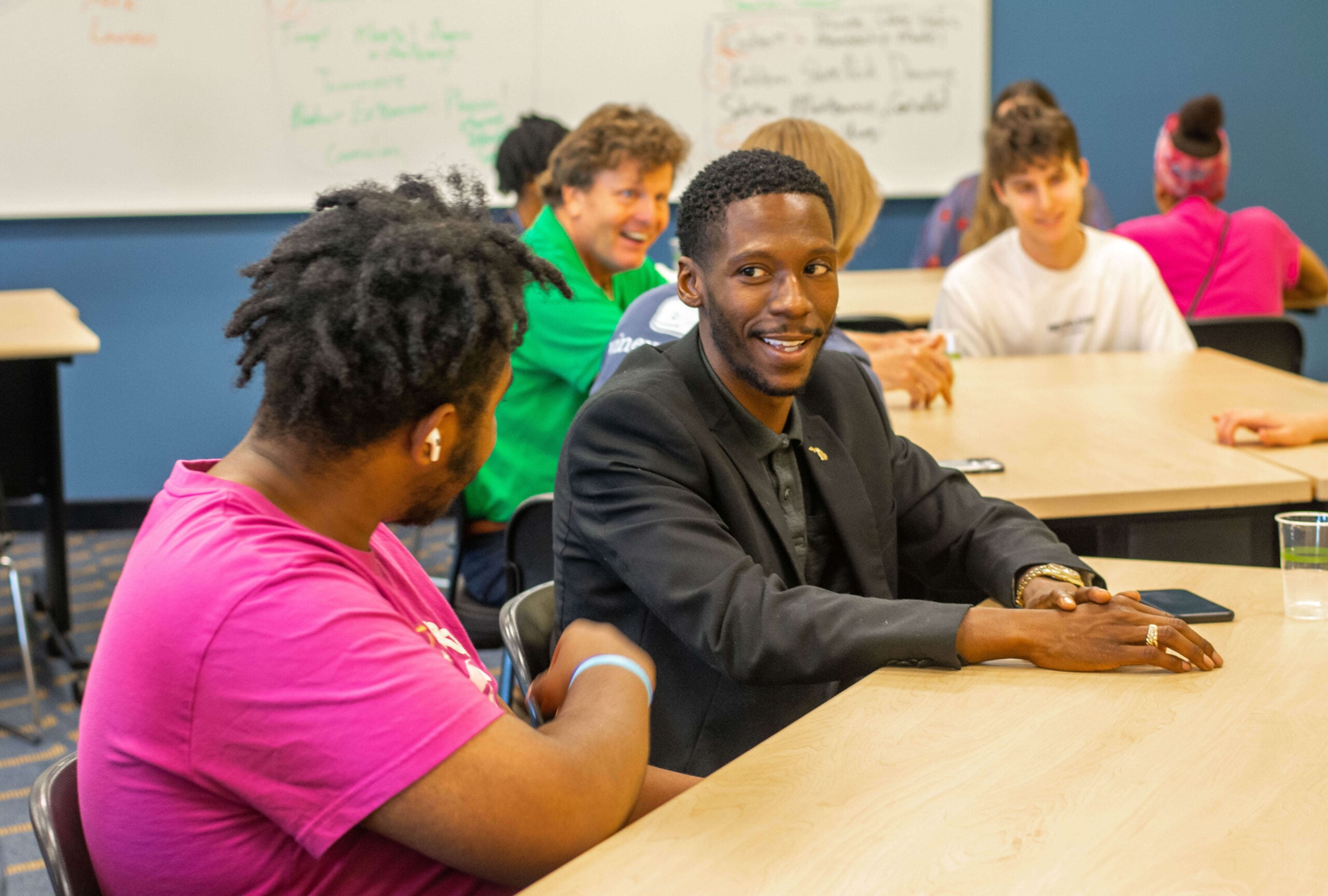 A business professional is seated at a table with a student, engaged in conversation. Others are smiling in the background.