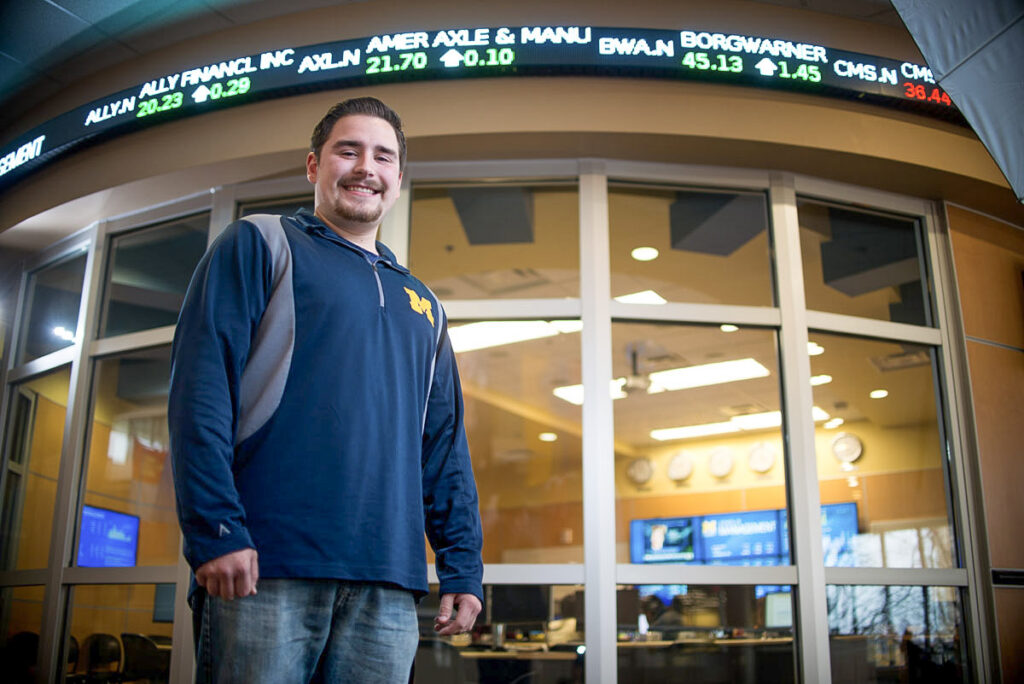 A young man smiles in front of a classroom's tall glass windows. This classroom is ringed with an electronic sign showing stock prices and market information.