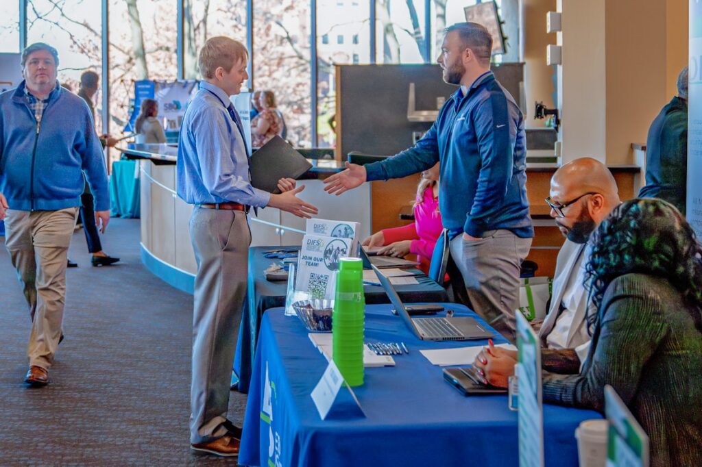 A man extends his hand across a table for a handshake during a career fair. A young man looks at his hand and prepares to shake it.