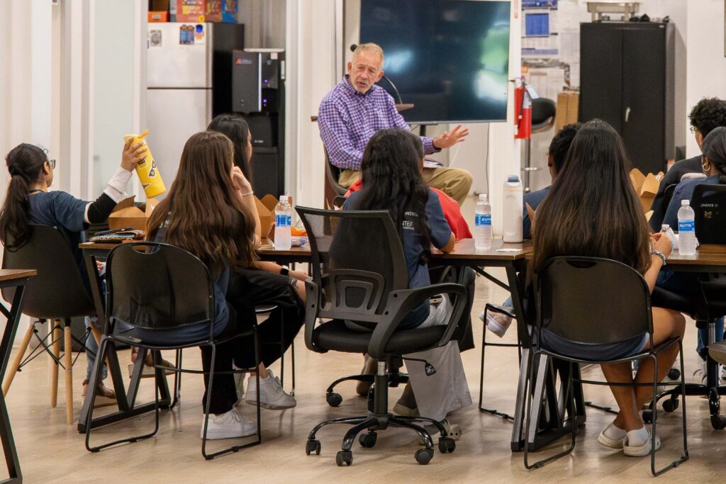A guest speaker talks to students seated at tables in a classroom setting. Each student is wearing a navy-blue University of Michigan-Flint t-shirt.