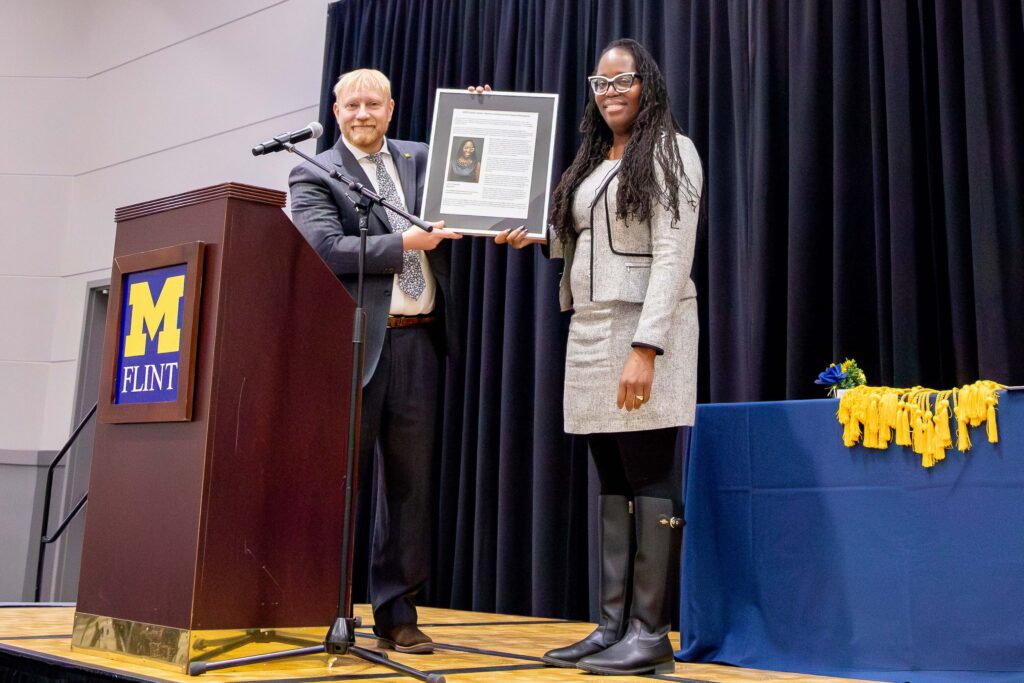A man presents a framed proclamation to a woman. Both are wearing business attire and standing at a podium displaying the University of Michigan-Flint logo.