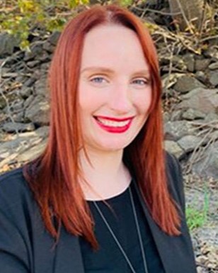 A woman with red hair and red lipstick smiles while standing outdoors near a rocky hillside. She is wearing black business attire.