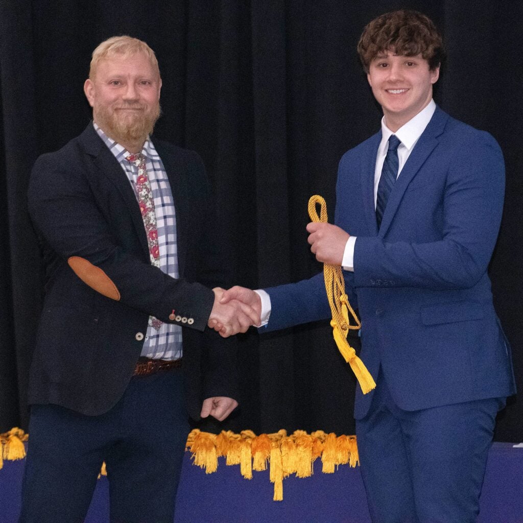 A professor shakes hands with a student during an Honors Night ceremony. The student is holding yellow honors cords.