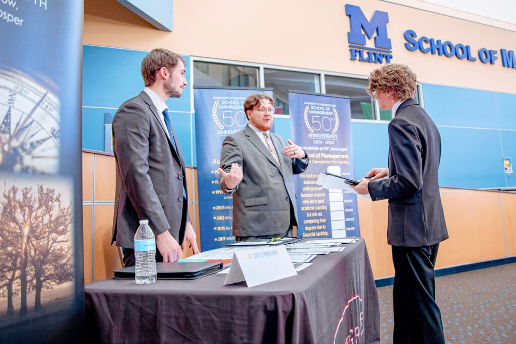 Two men are engaged in conversation with another man. They are all wearing gray business suits and standing at a table displaying information.