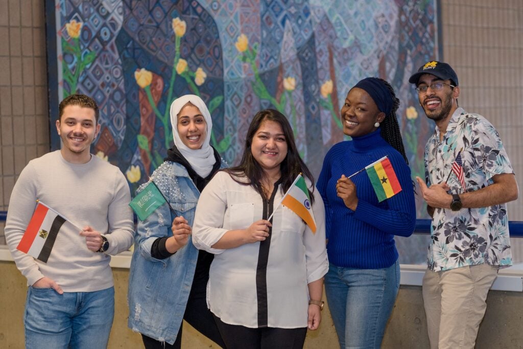 A group of five diverse people smile while holding flags from different countries. They are standing in front of a colorful mural.
