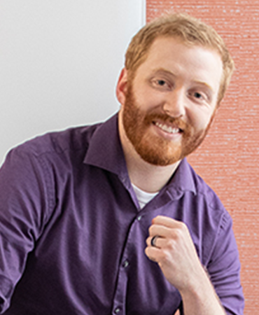 A professional portrait of a smiling man with a red beard and hair. He is wearing a purple, button-down shirt and a black ring on his left hand. The background is a vertical, woven tapestry that is light red on an off-white wall.