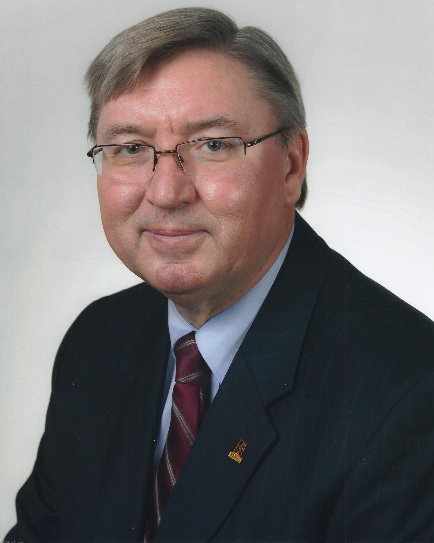A man in a navy-blue suit and maroon, striped tie is smiling at the camera. The background is a neutral gray.