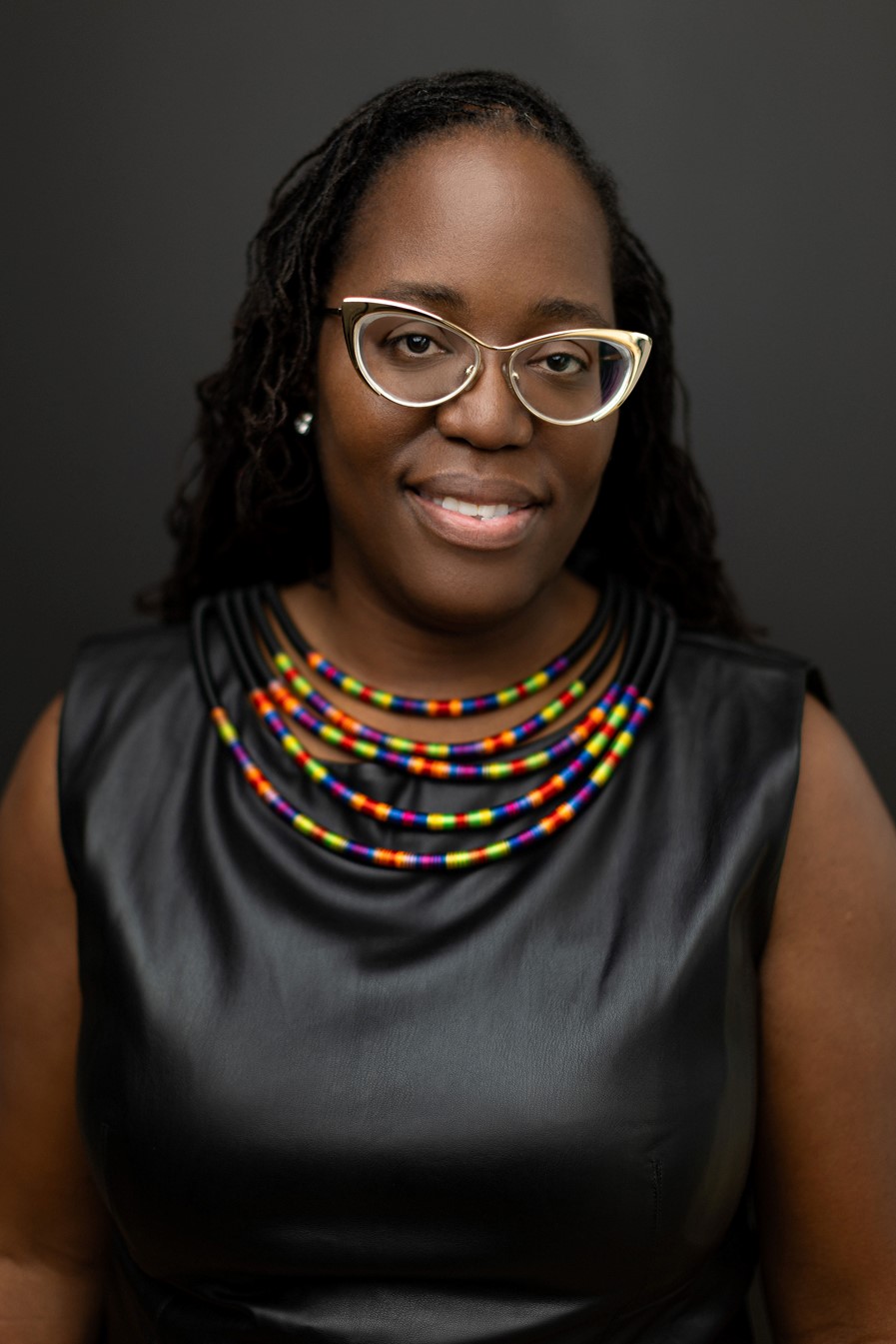 A woman with shoulder-length, black hair smiles against a dark background. She is wearing horn-rimmed glasses, a black leather top, and a necklace with colorful beads.