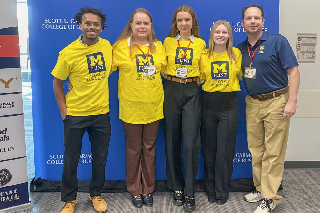 Four students and their professor stand in front of a blue backdrop. Each student is wearing a yellow t-shirt showing the University of Michigan-Flint logo.