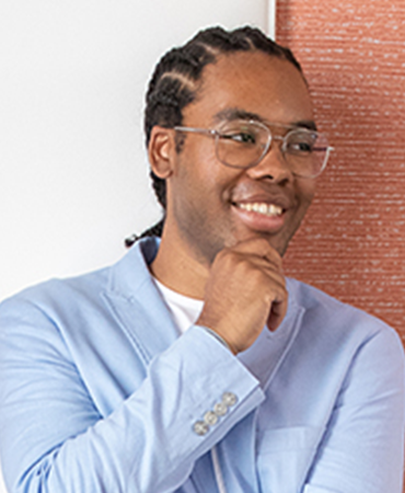 A professional portrait of a smiling man with short black braids. He is wearing round, wire-frame glasses and a powder-blue blazer. The background is a vertical, woven tapestry that is light red on an off-white wall.
