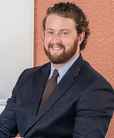 A professional portrait of a man with dark, curly hair and a beard. He is wearing a navy-blue suit and a solid, dark-brown tie. The background is a vertical, woven tapestry that is light red on an off-white wall.