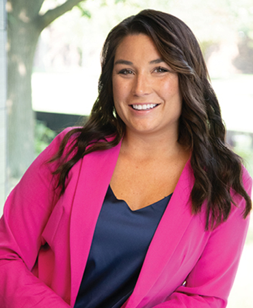 A portrait of a smiling woman with long brown hair. She is wearing a hot-pink blazer over a navy-blue top. She is standing indoors near a window overlooking a tree.